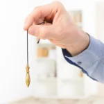 Una mano sostiene un péndulo dorado - curso de radiestesia